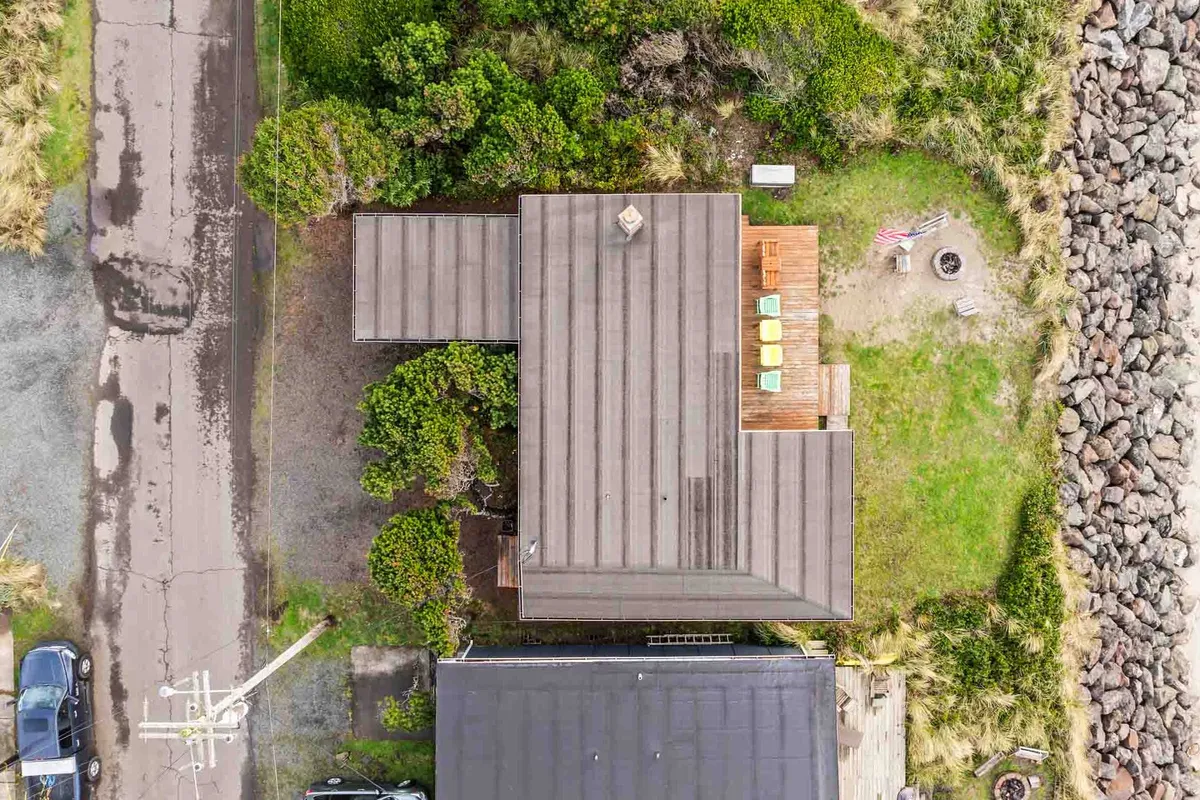 The image shows a broad perspective of the house's exterior, emphasizing the spacious deck area, a central fire pit, and surrounding greenery. The setup is ideal for barbecuing, relaxing outdoors, and gathering with loved ones as the sun sets, connecting indoor and outdoor living seamlessly.