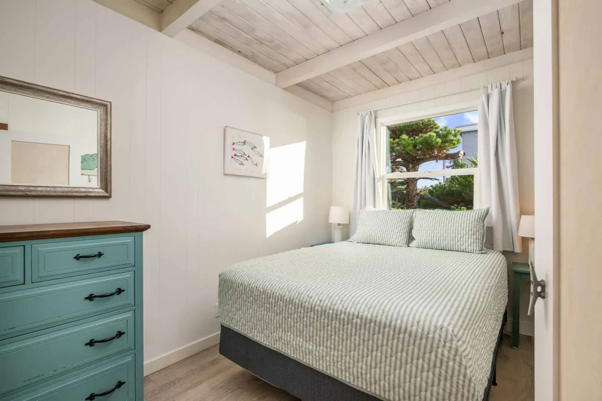 A restful bedroom scene featuring a neatly made bed, a window letting in gentle daylight, a dresser, tasteful artwork, and a nightstand. The space is airy and uncluttered, designed for quiet relaxation.