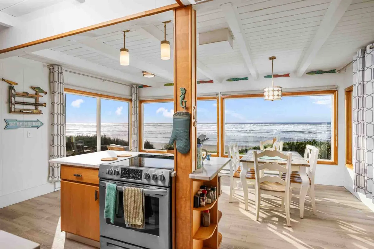 This image showcases a bright, welcoming kitchen with a kitchen island featuring a stove, a dining table surrounded by white chairs, and large windows that provide sweeping views of the beach and ocean. The setup suggests easy meal preparation and convivial family gatherings, with sunlight pouring in and the coastline visible beyond.