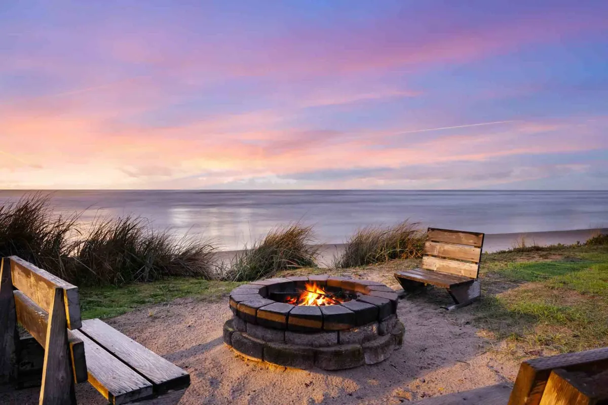 A cozy outdoor fire pit setup featuring wooden benches facing the beach, with golden sunlight casting a warm glow as the sun sets over the water. The scene is inviting for group gatherings, storytelling, and enjoying s'mores under the stars.