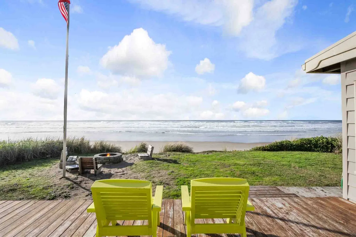The image showcases a cozy fire pit surrounded by cheerful yellow chairs on a patio overlooking the Pacific Ocean. A flag waves gently in the breeze, and the expansive ocean view stretches out to the horizon, with the beach just steps away. The scene is peaceful and inviting, perfect for sunset gatherings or stargazing by the fire.