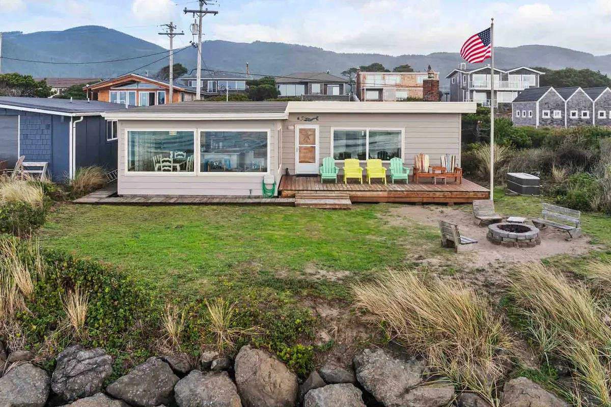 A cheerful deck scene with colorful chairs surrounding a fire pit, large windows reflecting the ocean, and the American flag waving in the background. The setting is primed for socializing, cooking out, or simply soaking in ocean breezes and panoramic views.