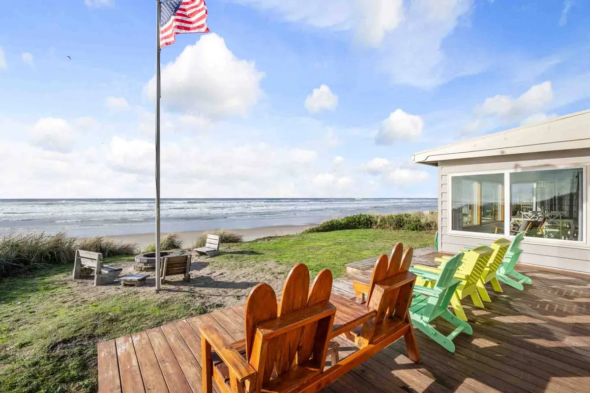 An inviting outdoor fire pit area is surrounded by colorful Adirondack chairs, all facing the ocean. The scene captures the sun's glow over the water, with the deck and firepit positioned to maximize enjoyment of the seaside atmosphere. The image hints at both al fresco dining and fireside relaxation, with a sense of warmth and camaraderie.