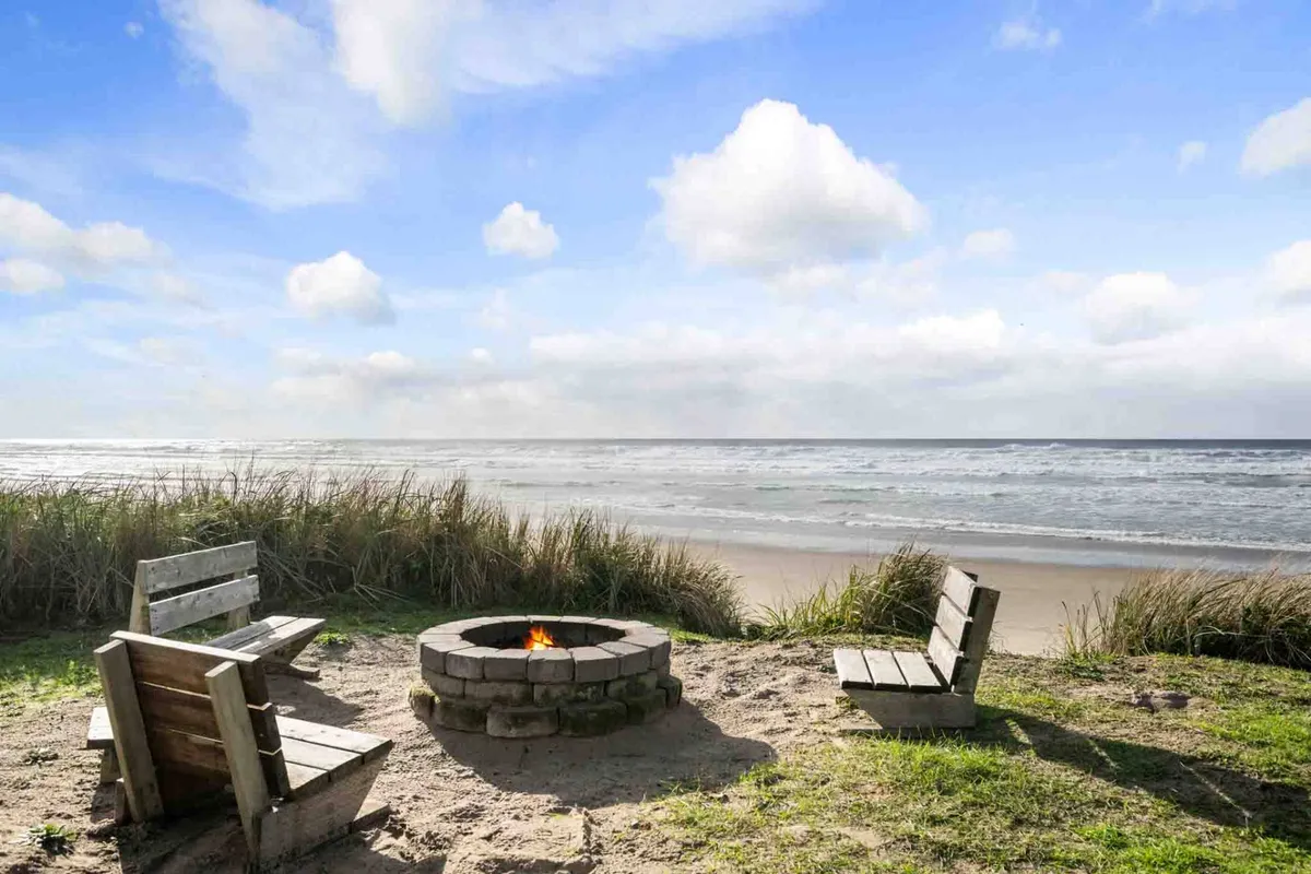 This image features a cozy fire pit setup on a deck overlooking the beach and ocean, with comfortable seating arranged for group gatherings. The sky glows with warm colors as the sun sets, creating a relaxing and inviting atmosphere. The fire pit is central, suggesting warmth and togetherness, and the sandy beach is just steps away.