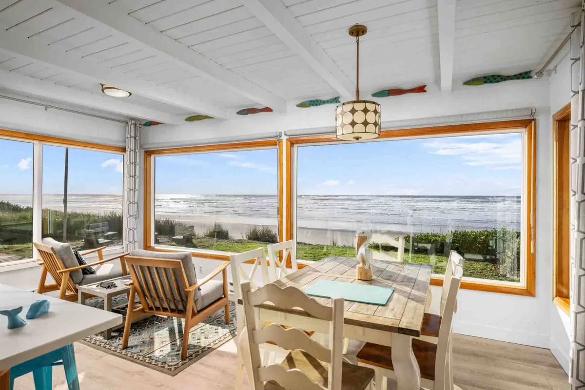 A welcoming interior scene with large picture windows framing spectacular ocean views. The image features a dining table set for a family or group, a comfortable seating area with plush chairs, and warm, natural light flooding in, creating a relaxed and inviting atmosphere.