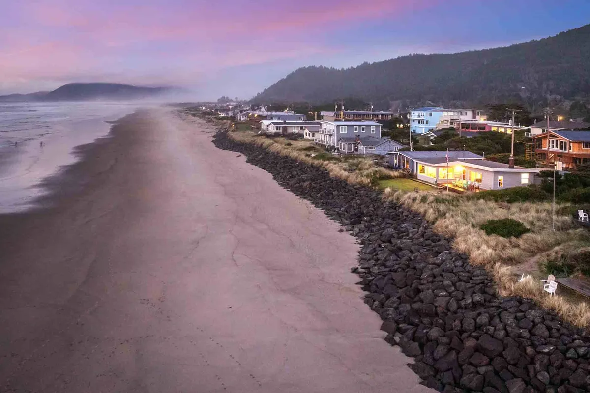 A wide-angle shot of beachfront houses facing a rocky shoreline, with waves crashing and a dramatic sunset sky overhead. The ambiance is tranquil and perfect for evening walks or group gatherings. The shoreline and open space make it ideal for exploring or simply enjoying the views together.