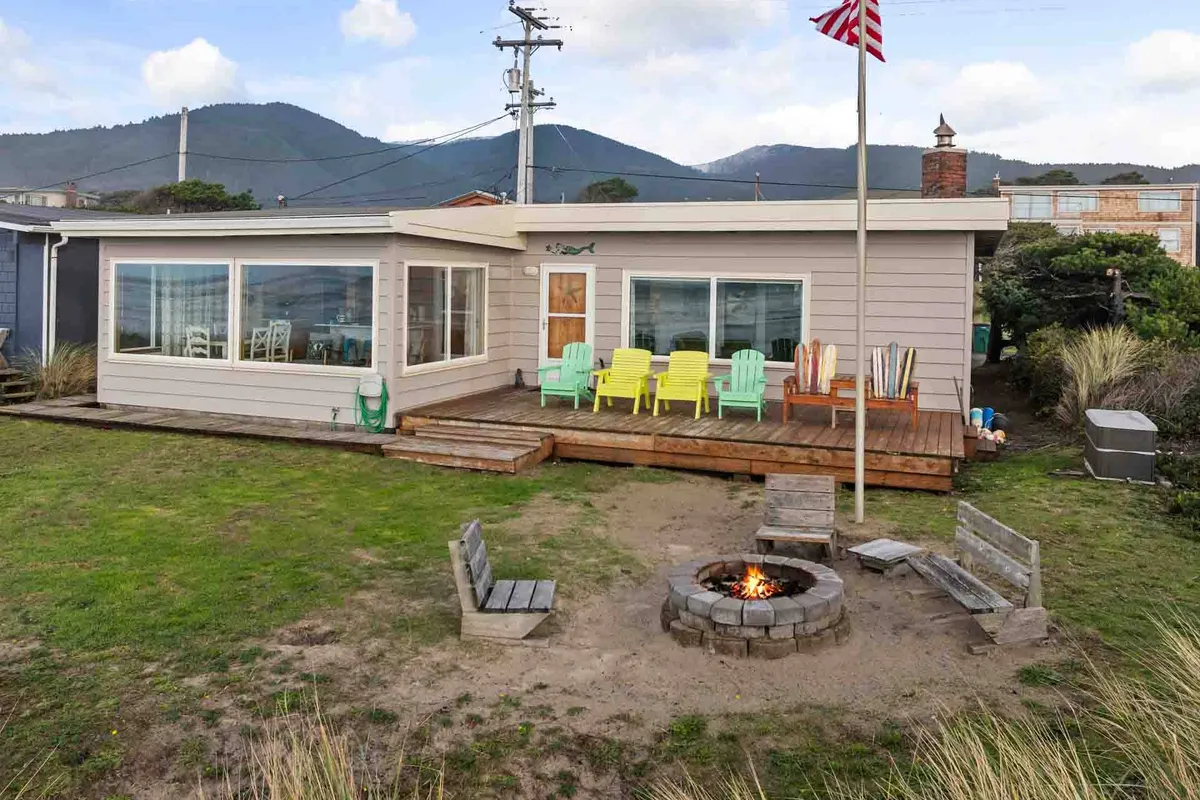 An inviting outdoor gathering space features colorful chairs arranged around a fire pit, with the home's large windows and a scenic mountain backdrop visible. The setting suggests relaxation and socializing in the fresh air.