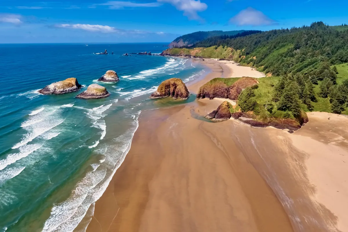 A sweeping view of the beach with striking rock formations rising from the sand, gentle waves lapping the shore, and a backdrop of dense coastal forest under a serene sky. The warm hues of sunset cast a romantic glow over the entire landscape, inviting leisurely strolls along the water's edge.
