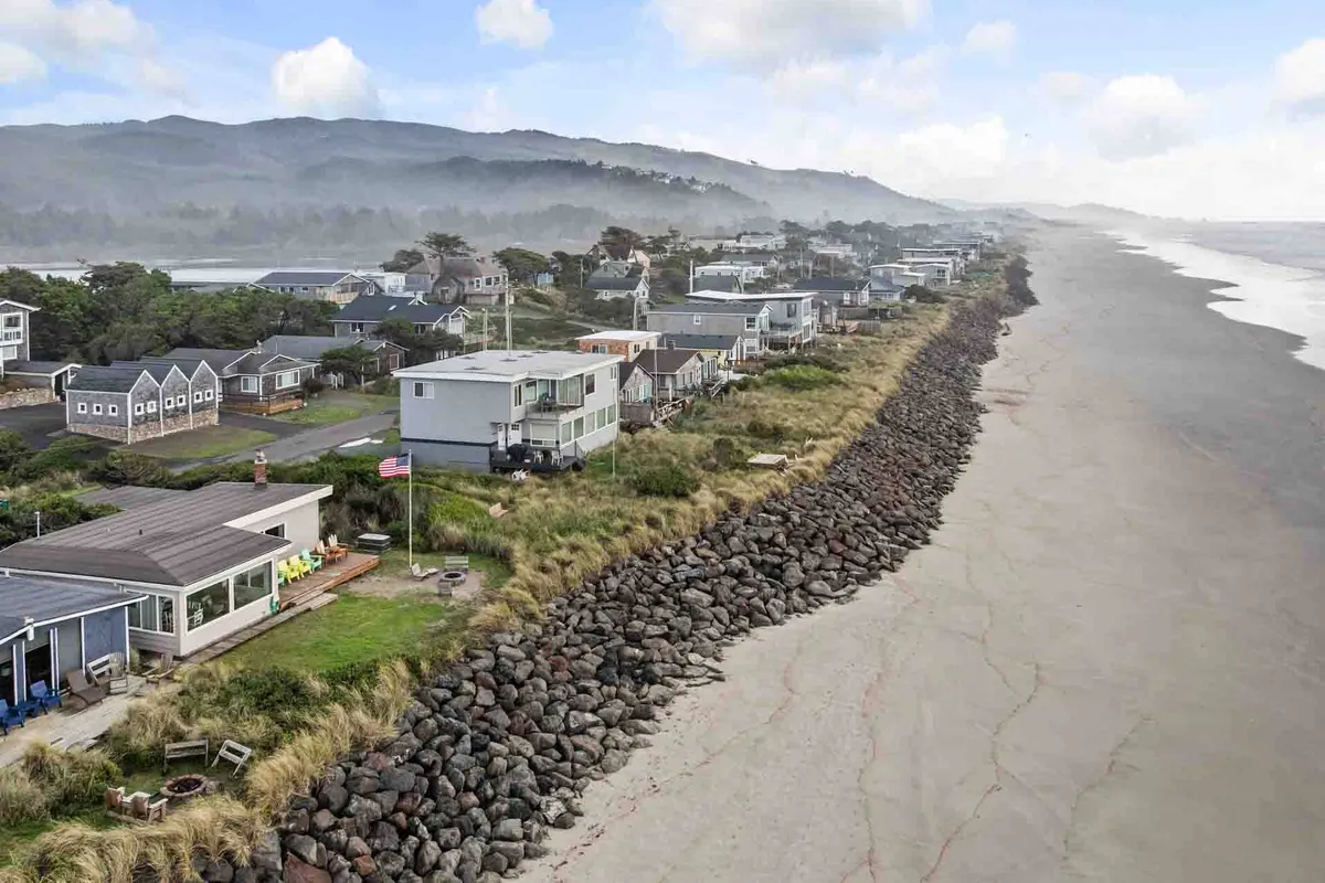 A serene stretch of sandy beach bordered by rocky shoreline and beachfront houses, with distant hills in view. The image captures a tranquil scene where children could easily be imagined building sandcastles while families enjoy strolls along the water's edge.