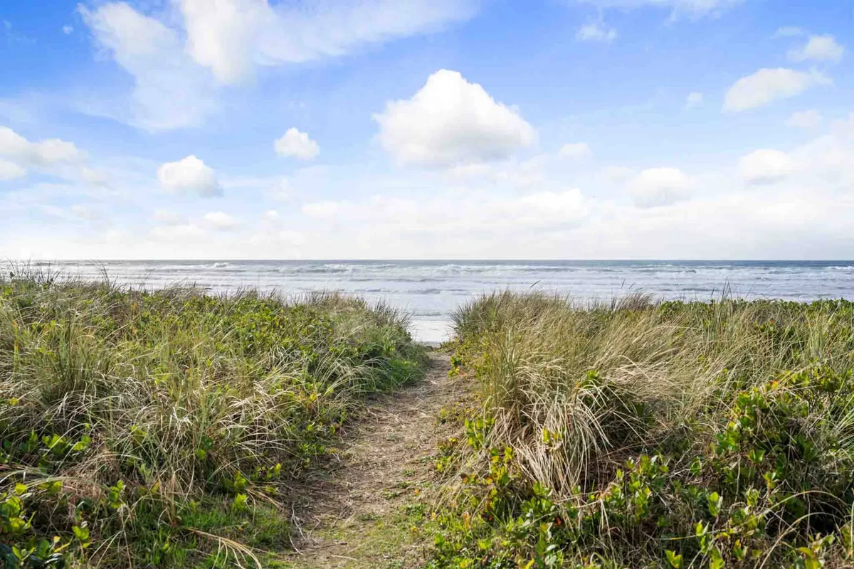 A tranquil view along a sandy pathway through dune grass leading directly to the ocean, framed by open sky. The natural, unspoiled environment invites peaceful strolls and direct access to the beach.