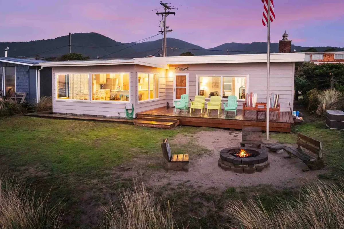 An inviting outdoor space featuring a fire pit surrounded by comfortable outdoor seating, large windows reflecting nature, and a flagpole, all set against a tranquil backdrop. The area is ideal for gatherings after a day at the beach, with plenty of room for pets and owners to unwind together.
