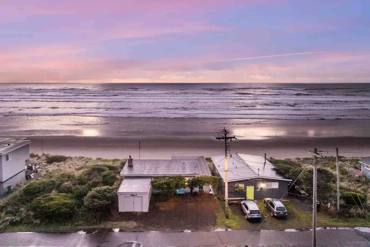 This exterior image shows the beachfront house set against a stunning sunset sky, with ocean waves rolling in. Several cars are visible parked in front of the house, while power lines and the natural shoreline emphasize the property's coastal location.