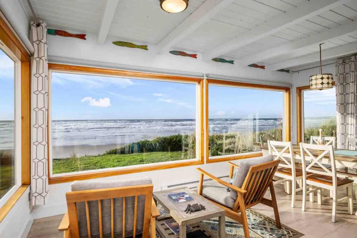 A tranquil dining area with large windows overlooking the ocean, wooden chairs around a welcoming table, and soft, golden light. The setup invites family gatherings, board games, and shared meals, all while taking in beautiful coastal views.