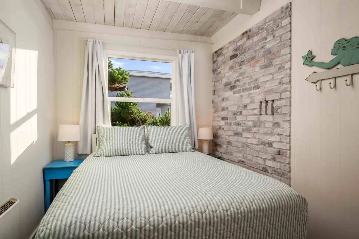 A serene bedroom with a bed dressed in a patterned quilt, a window with soft curtains letting in natural light, a textured brick accent wall, and a blue side table. The room feels restful and inviting, ideal for unwinding after a day at the coast.